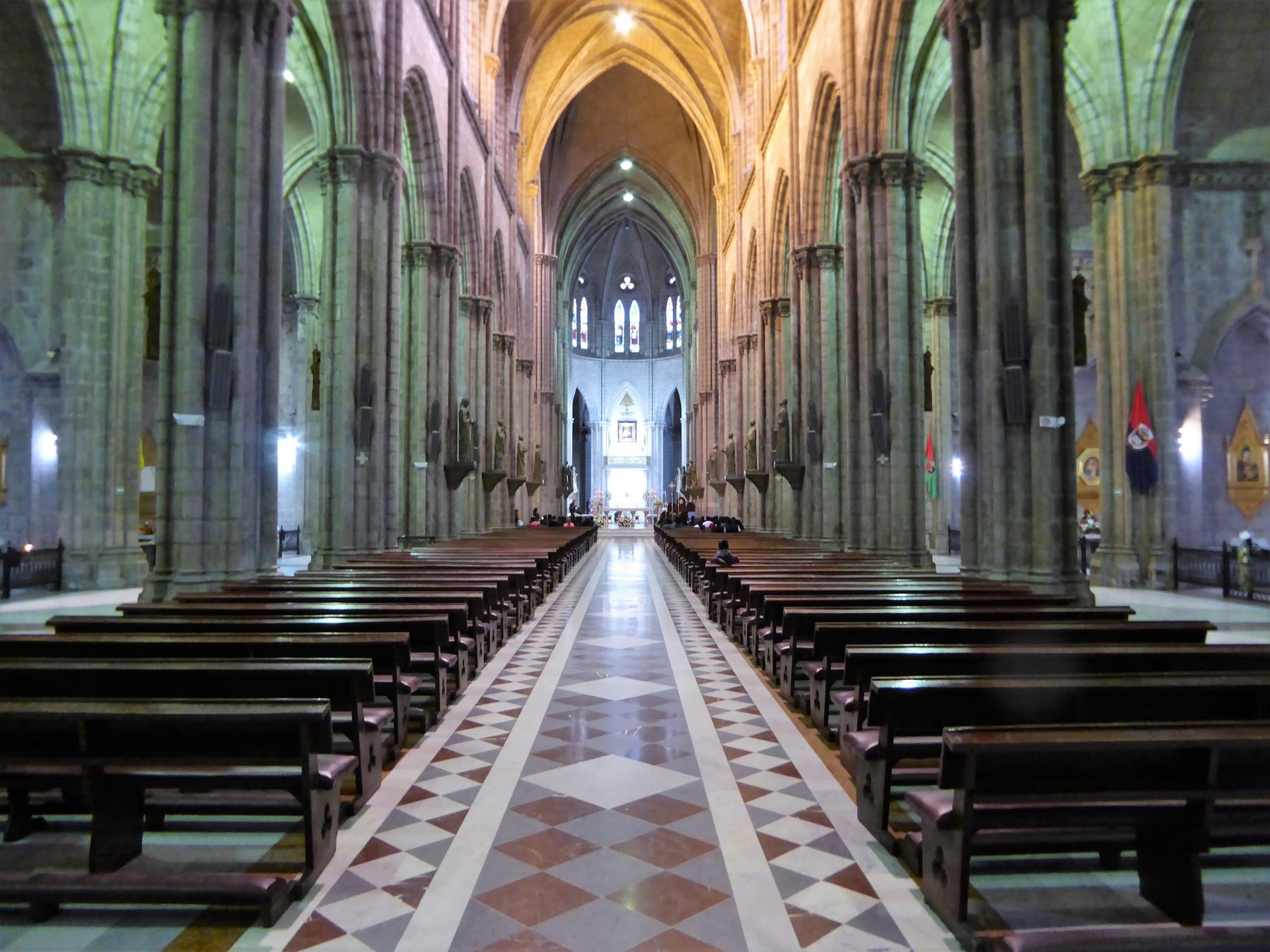View down the center aisle of the Basílica del Voto Nacional from the back of the nave, framed by stone columns and rows of wooden pews leading to the high altar in the distance