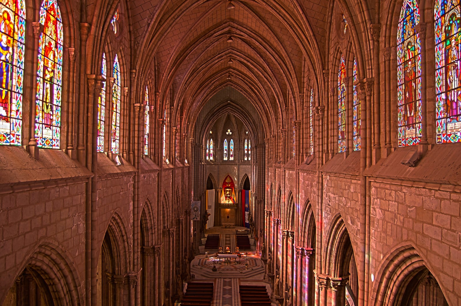 The Gothic nave of the Basílica del Voto Nacional seen from the choir loft, with soaring red-stone arches and stained-glass windows on both sides leading to the altar