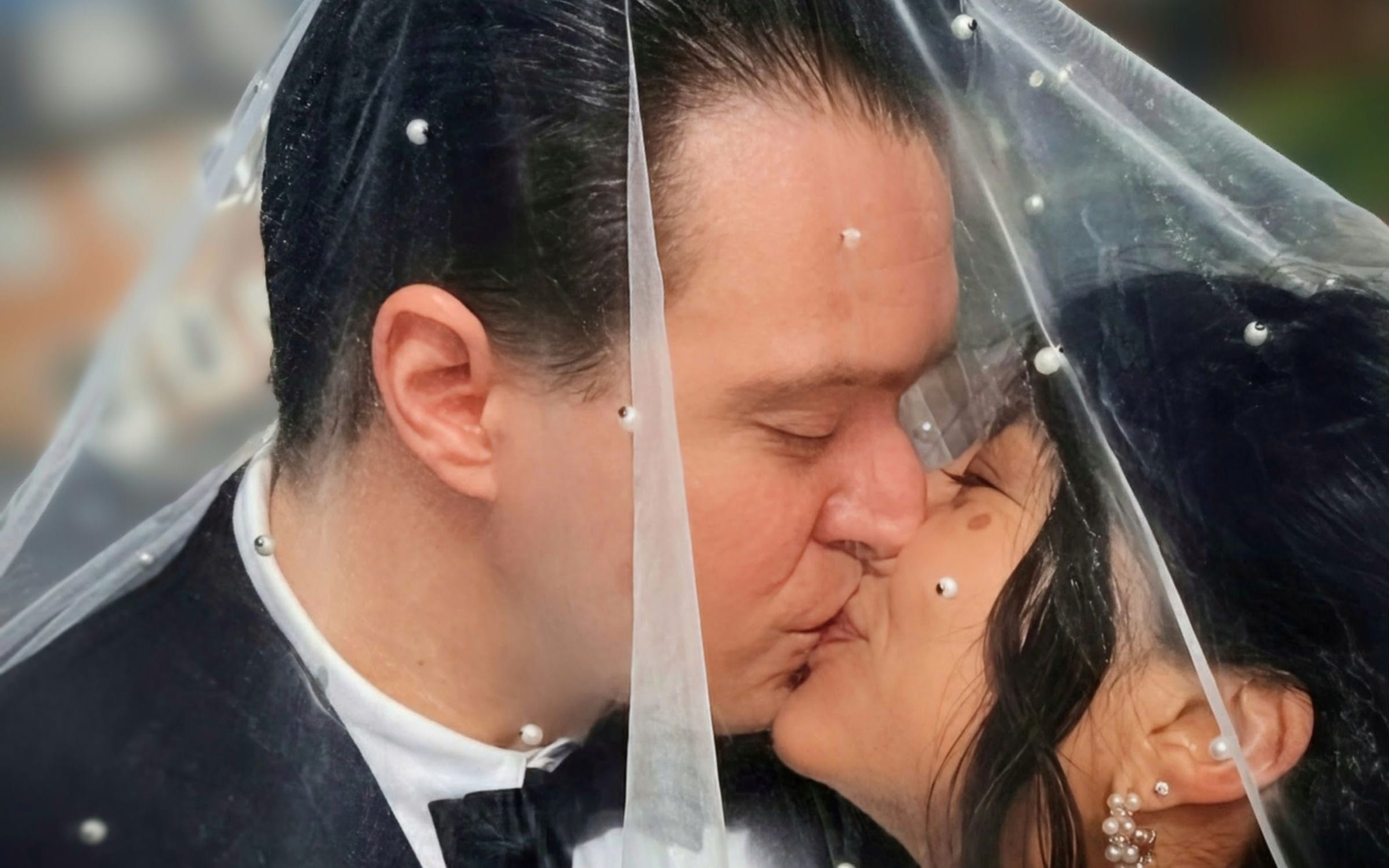 John and Diana sharing a kiss beneath a pearl-studded veil, with city buildings and blue sky behind them