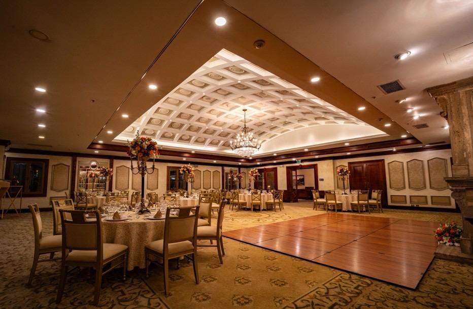 The Hotel Plaza Grande ballroom with round tables, gold candelabras with floral arrangements, and a wooden dance floor
